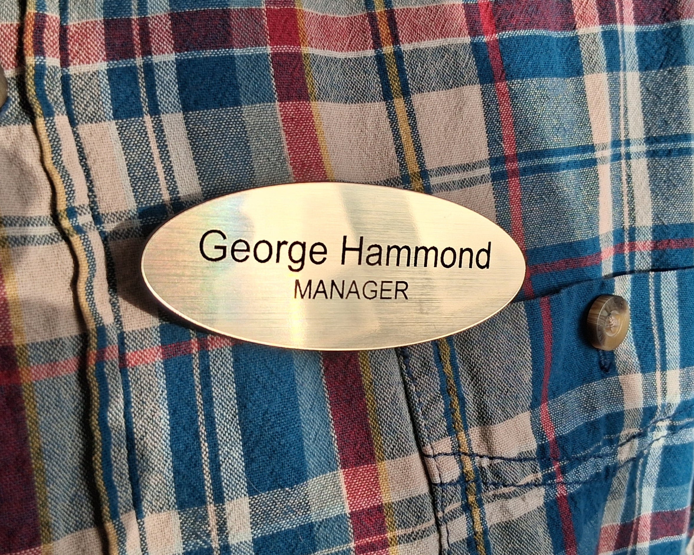 Plaid shirt with a custom engraved plastic (lamacoid) name tag reading 'George Hammond Manager' on a dark background