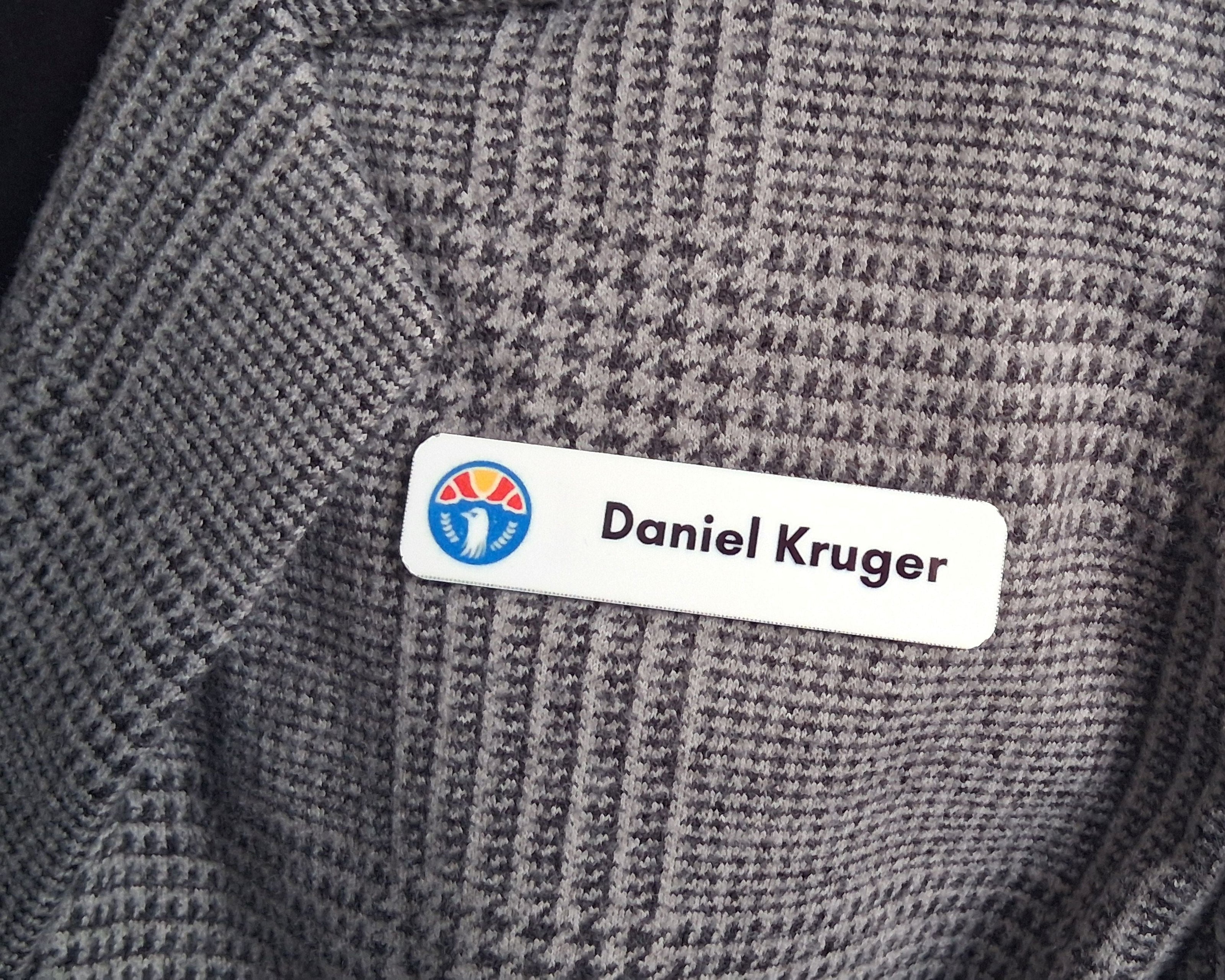 Gray textured jacket with a custom metal full colour name tag labeled 'Daniel Kruger'.
